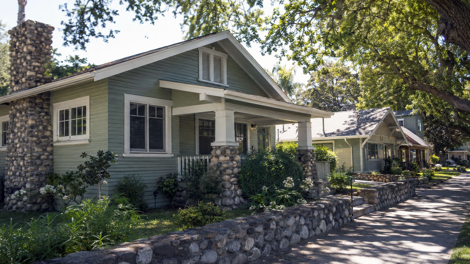 Sage green Craftsman bungalow in Claremont, river rock chimney and porch piers, a low rock wall along the sidewalk.